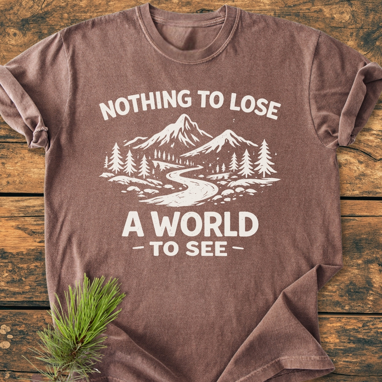 Vintage Wash Tee with a white graphic of a river, mountains, and pine trees, phrases “NOTHING TO LOSE” and “A WORLD TO SEE,” laid flat on wood with a pine sprig.