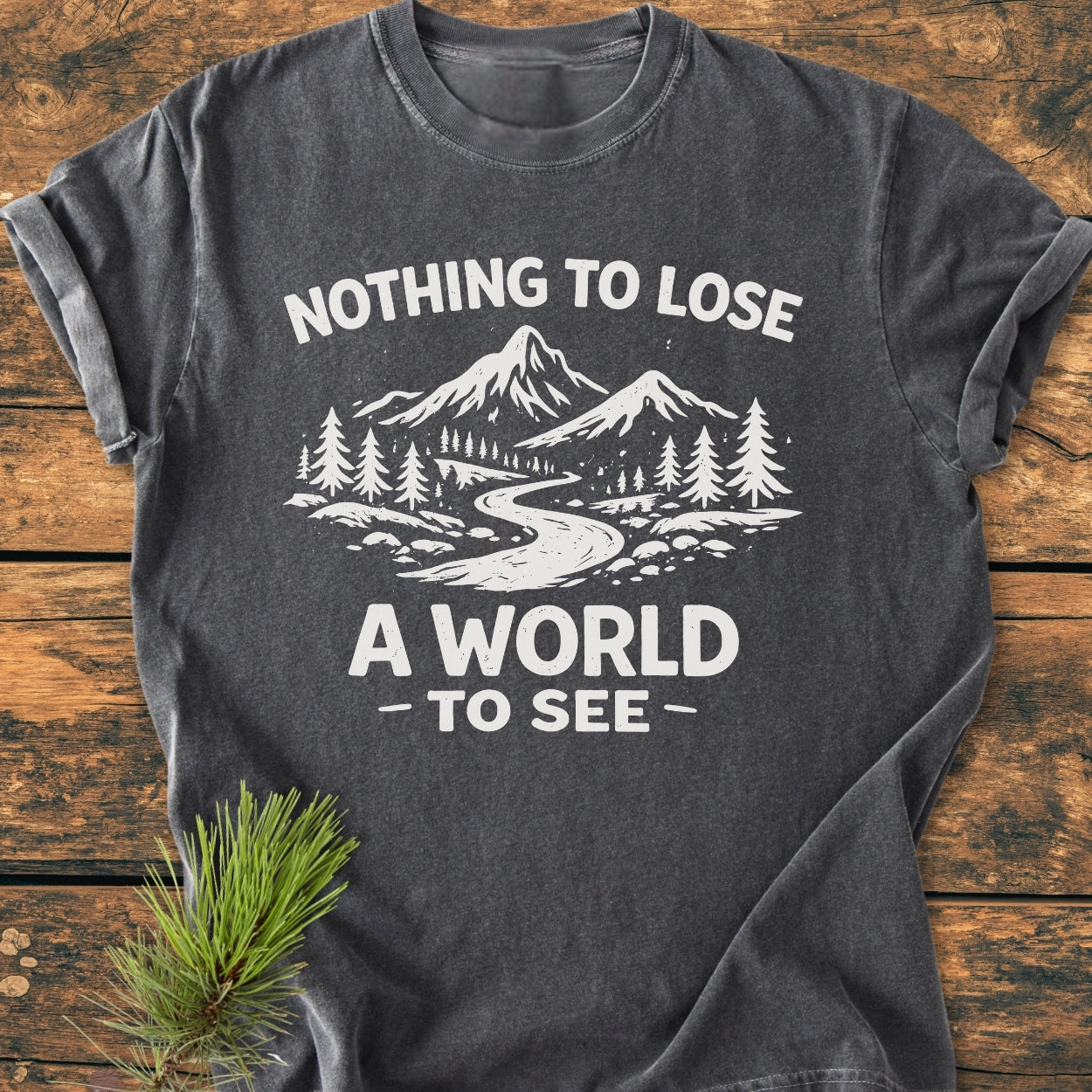Nothing To Lose - Vintage Wash Tee features a white outdoors graphic of mountains and a trail on a charcoal-grey shirt, laid on wood with a pine sprig, showcasing its cozy, relaxed fit.