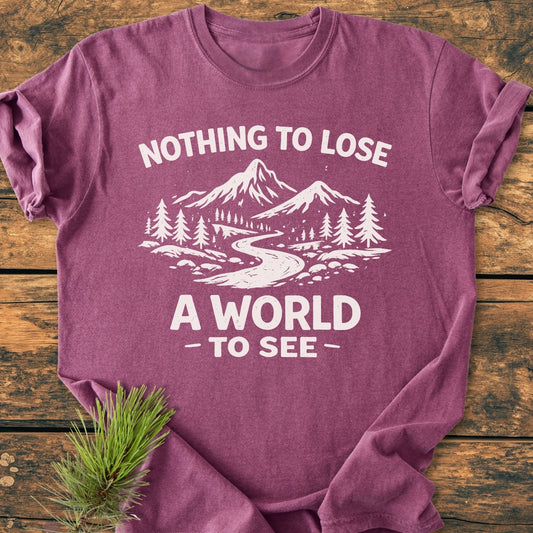 Nothing To Lose - Vintage Wash Tee features a white screen-printed mountain scene with NOTHING TO LOSE and A WORLD TO SEE slogans, folded on a rustic wooden surface with pine sprig accent.