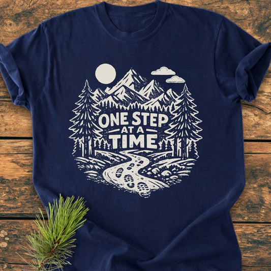 One Step At A Time - Vintage Wash Tee features a white, hand-drawn outdoor scene with pine trees, mountains, and a motivational phrase, printed on a navy-blue, soft-washed, 100% cotton T-shirt.