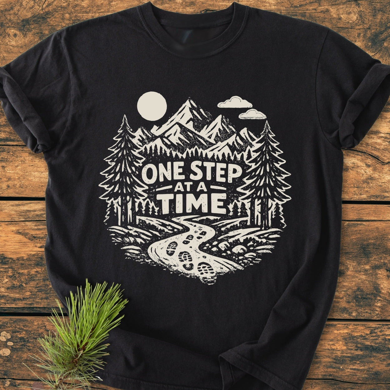 One Step At A Time - Vintage Wash Tee displayed flat, featuring a stylized outdoors scene with a trail, pine trees, mountains, and bold lettering on soft, garment-dyed cotton for a cozy, relaxed fit.