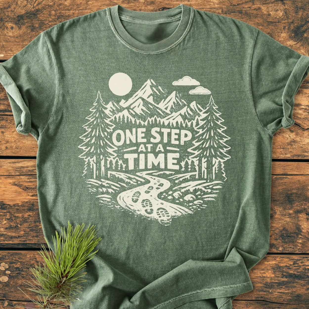 One Step At A Time - Vintage Wash Tee features a heather-green T-shirt with a mountain range, pine trees, and trail graphic, plus a motivational slogan, offering a cozy and durable relaxed fit.