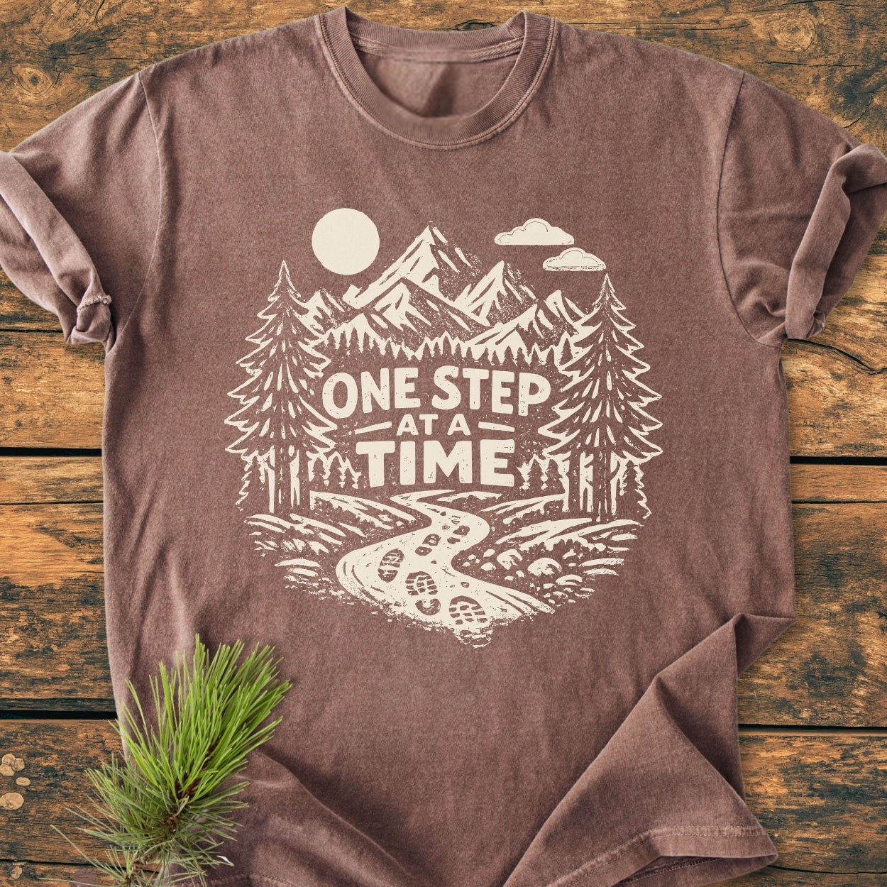 One Step At A Time - Vintage Wash Tee features a stylized outdoor scene with mountains, trees, and a trail, screen-printed in off-white on a brown crew-neck t-shirt, laid flat on wood.