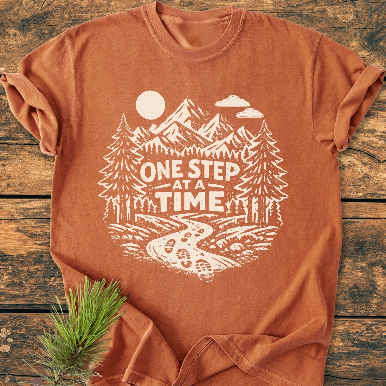 One Step At A Time - Vintage Wash Tee: Rust-orange cotton T-shirt featuring white outdoor-themed graphic with pine trees, mountains, and trail. Laid on a wooden surface with a pine branch beside it.