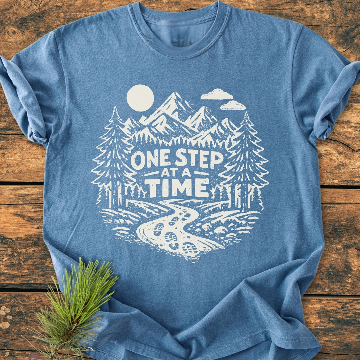 One Step At A Time - Vintage Wash Tee features a heather-blue backdrop with a white print of a mountain trail scene and slogan, highlighting its relaxed fit and nature-inspired design.