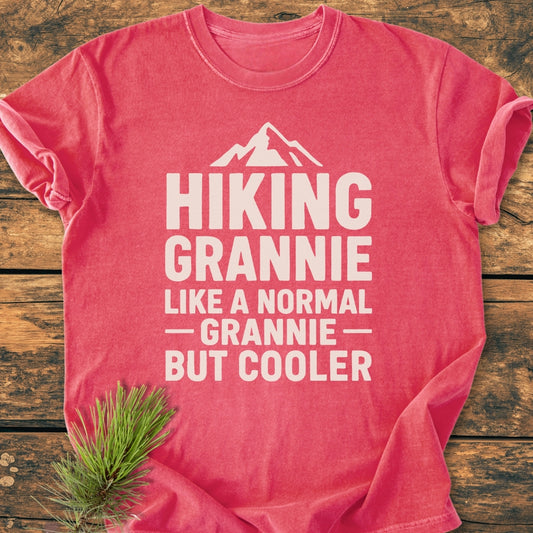 The image displays the Hiking Grannie - Vintage Wash Tee, a coral-red T-shirt with a mountain icon and slogan, styled on a wooden surface with a pine sprig.