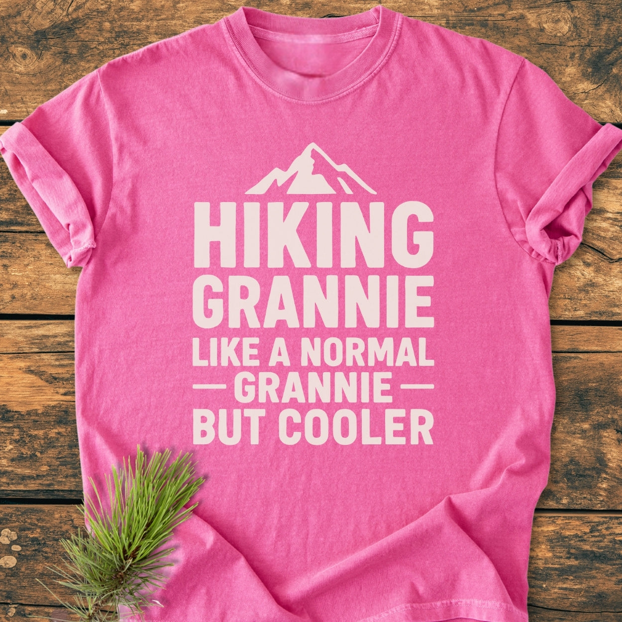Hiking Grannie - Vintage Wash Tee: A bright pink T-shirt with bold white lettering and mountain graphic. Laid on wood with pine sprig, showcasing cozy, relaxed fit and durable, ring-spun cotton fabric.
