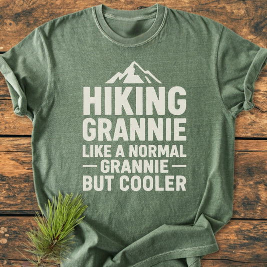 Hiking Grannie - Vintage Wash Tee: A heather-green T-shirt with HIKING GRANNIE slogan and mountain silhouette, laid on wood. Features soft, durable 100% ring-spun cotton, relaxed fit, decorative pine sprig accent.
