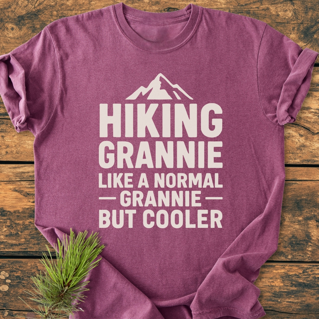 Hiking Grannie - Vintage Wash Tee features a purple-mauve T-shirt with a mountain silhouette and slogan, displayed on a wood tabletop with a pine bough, highlighting its relaxed, durable design.