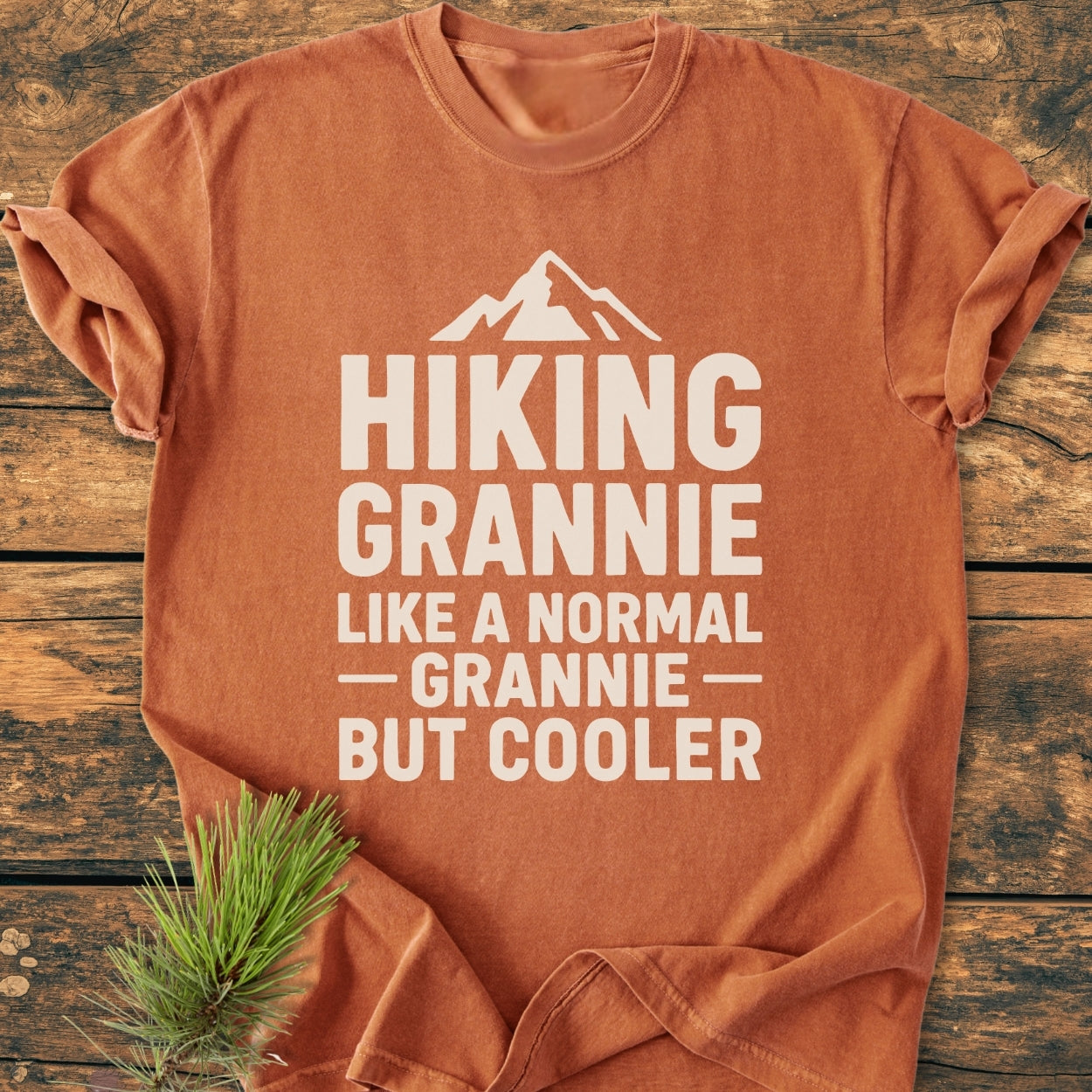 Hiking Grannie - Vintage Wash Tee: An orange-rust T-shirt with a white mountain graphic and playful text, laid on wood, accented by pine needles. Made from soft, durable, pre-shrunk ring-spun cotton.