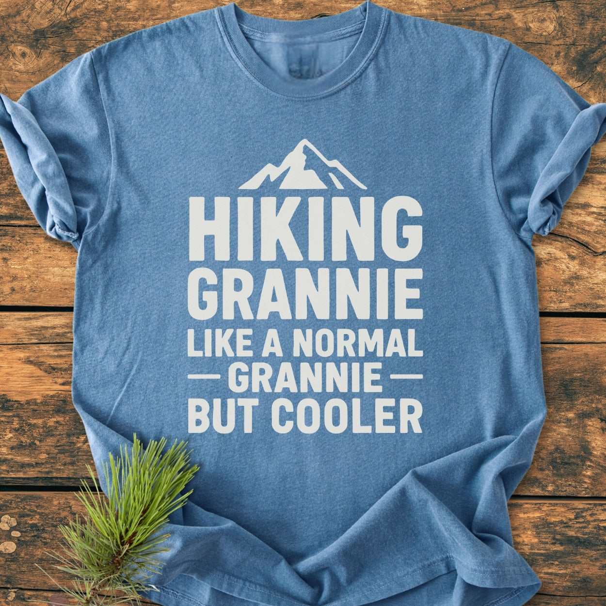 Hiking Grannie - Vintage Wash Tee featuring a mountain graphic and slogan, laid on rustic wood with a pine sprig. Made of soft, durable, garment-dyed, 100% ring-spun cotton for a relaxed fit.