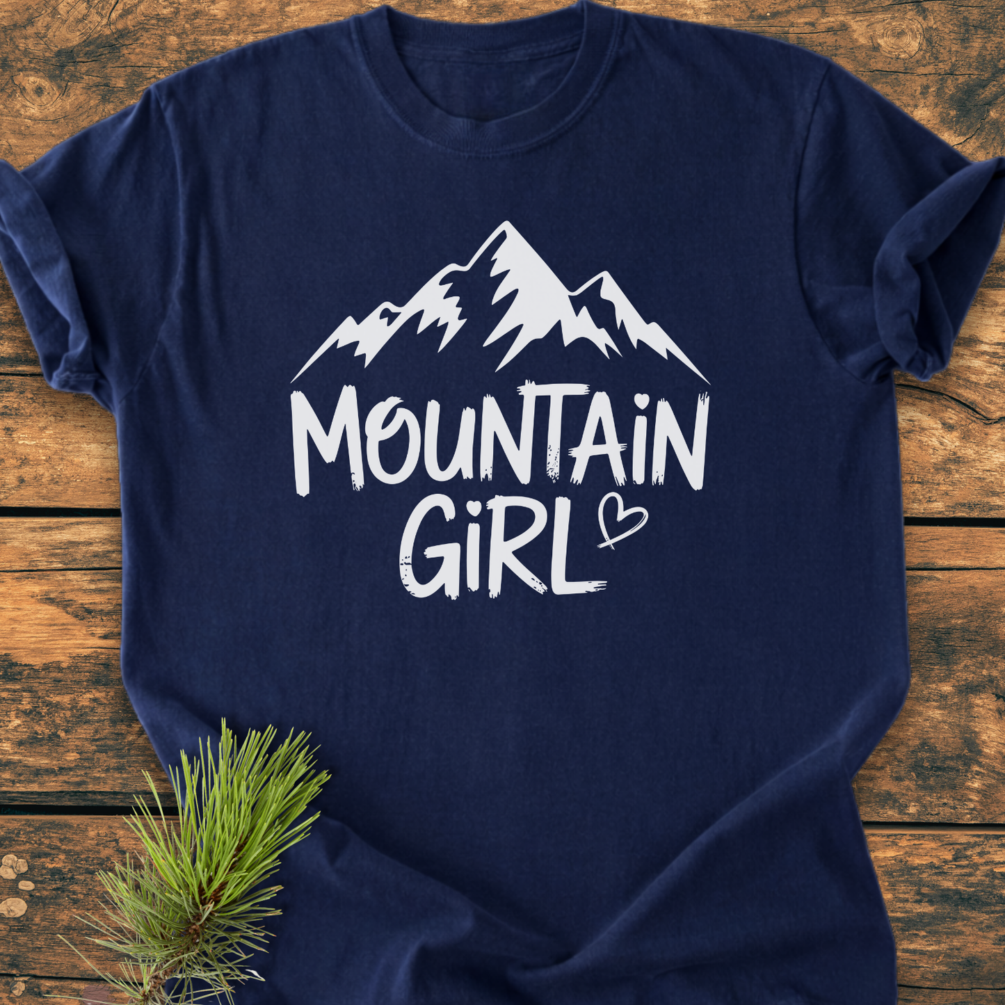Mountain Girl - Vintage Wash Tee features a navy-blue fabric with a stylized mountain range and MOUNTAIN GIRL in white. A small heart detail and a pine branch are also visible.