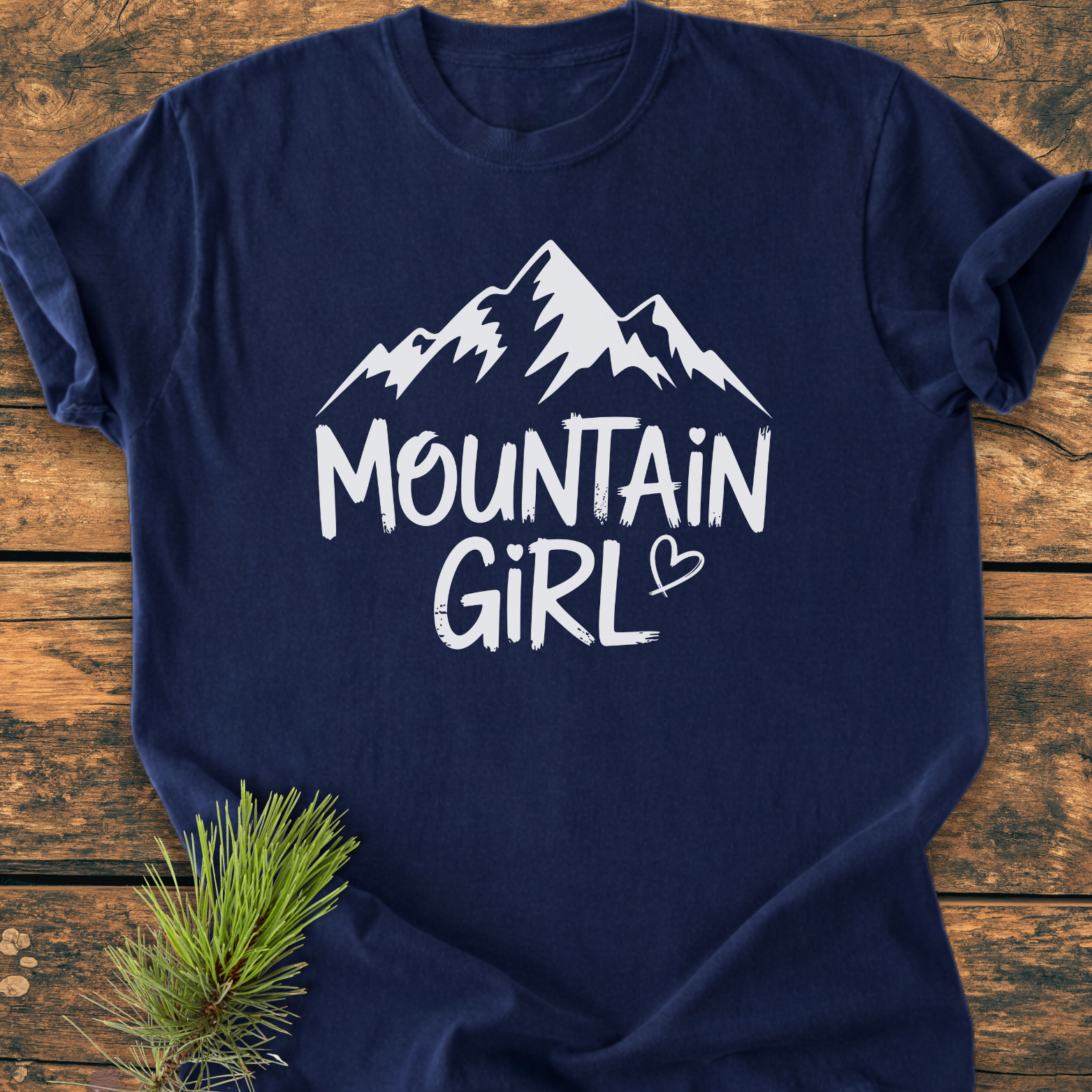 Mountain Girl - Vintage Wash Tee features a navy-blue fabric with a stylized mountain range and MOUNTAIN GIRL in white. A small heart detail and a pine branch are also visible.