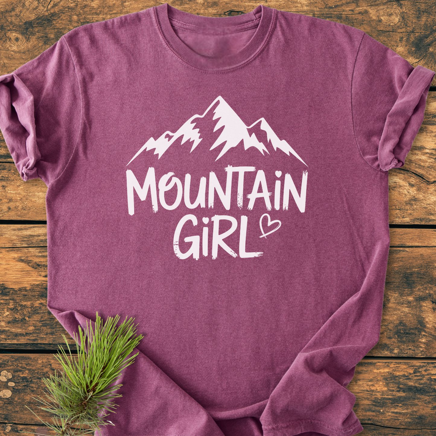 Mountain Girl - Vintage Wash Tee featuring a white mountain graphic and heart-dotted i in GIRL, laid on wood with an evergreen sprig, showcasing its soft, relaxed fit and durability.