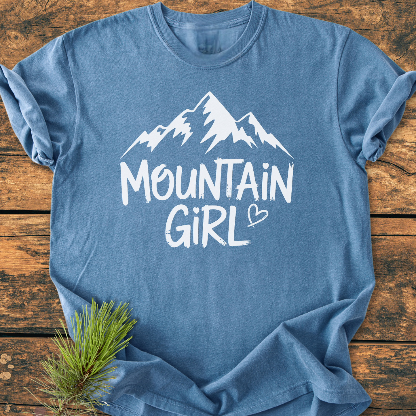 Mountain Girl - Vintage Wash Tee: Soft-blue crewneck T-shirt featuring a white mountain silhouette and MOUNTAIN GIRL text with a heart, laid on wood with a pine branch accent.