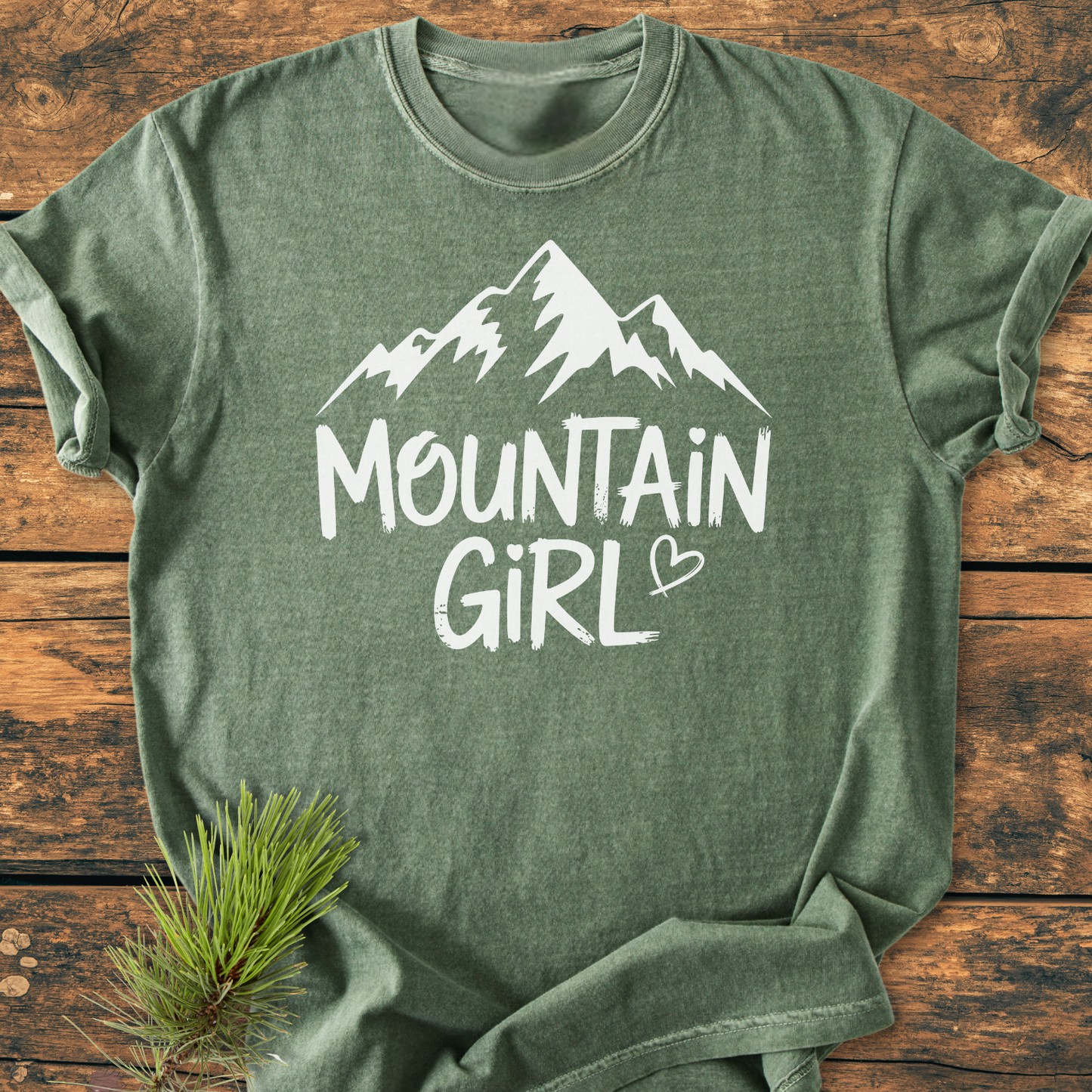 Mountain Girl - Vintage Wash Tee displayed flat, featuring a stylized mountain graphic and text. Tucked pine branch adds rustic charm, highlighting the shirt's relaxed, durable design.