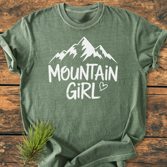 Mountain Girl - Vintage Wash Tee displayed flat, featuring a stylized mountain graphic and text. Tucked pine branch adds rustic charm, highlighting the shirt's relaxed, durable design.