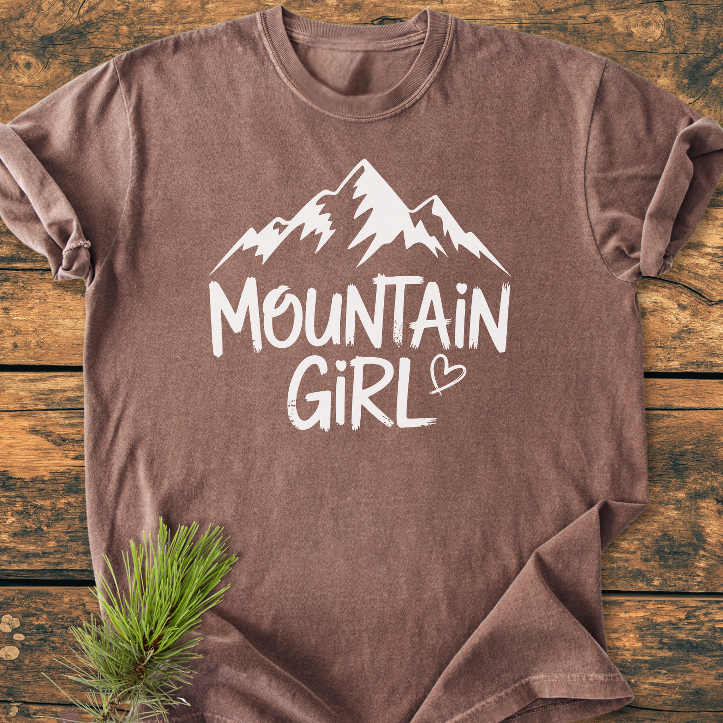 Mountain Girl - Vintage Wash Tee: Brown heathered T-shirt with a white mountain silhouette and MOUNTAIN GIRL text, laid flat on wood. Pine sprig accentuates the rustic presentation.