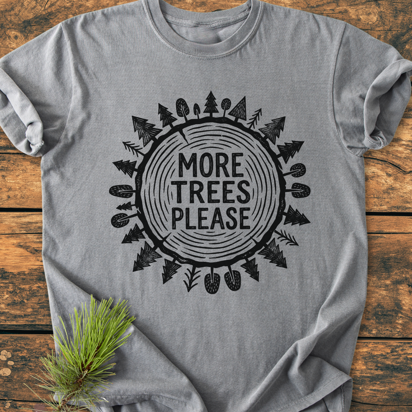 Heather-gray More Trees Please - Vintage Wash Tee with a tree-ring graphic and text, laid flat on wood, featuring durable, relaxed fit, made from 100% ring-spun cotton, with a pine needle accent.