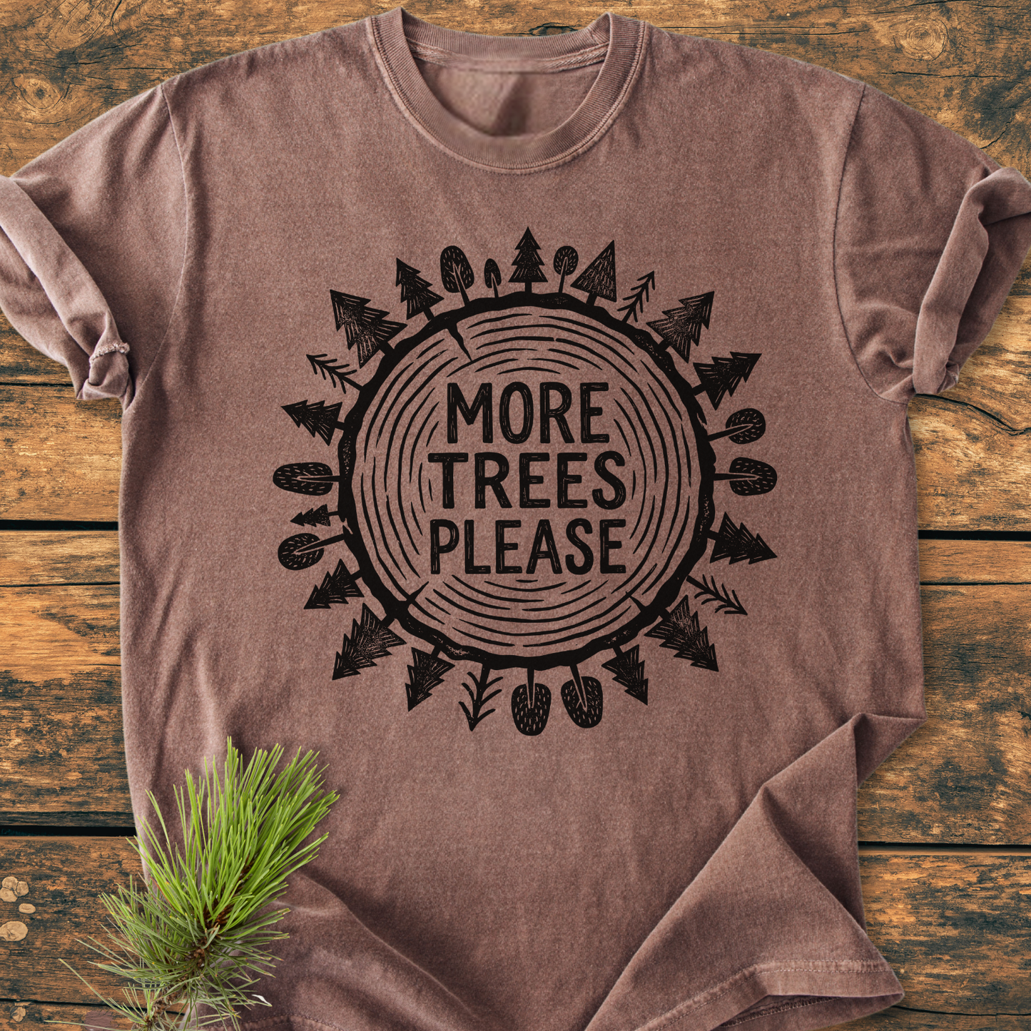 More Trees Please - Vintage Wash Tee features a heathered brown design with a circular tree ring graphic and nature motifs. Displayed on wood, it embodies comfort with its relaxed fit and durable stitching.