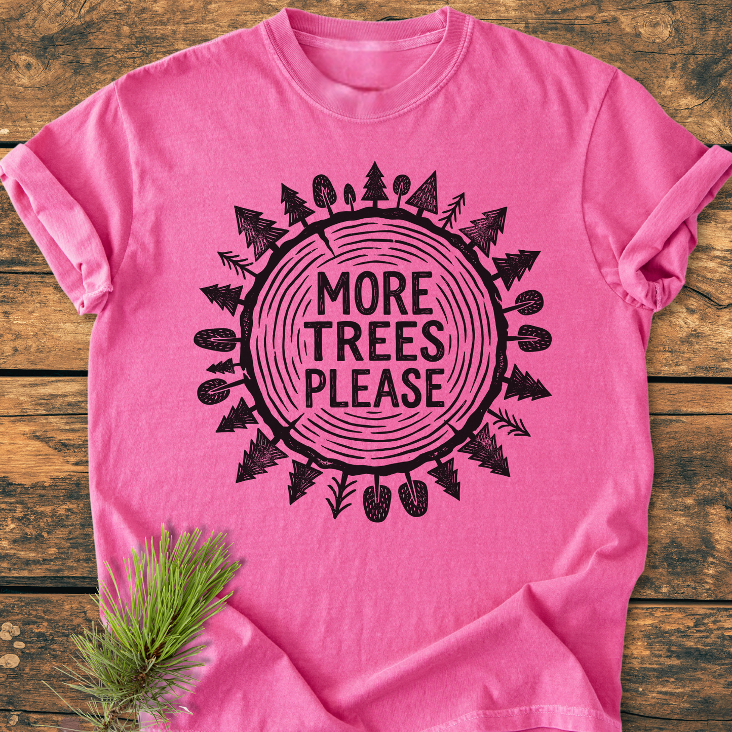 More Trees Please - Vintage Wash Tee displayed on wooden surface, featuring a tree-ring graphic and slogan. Pine sprig beside it. Soft, relaxed fit made from 100% ring-spun cotton enhances comfort.