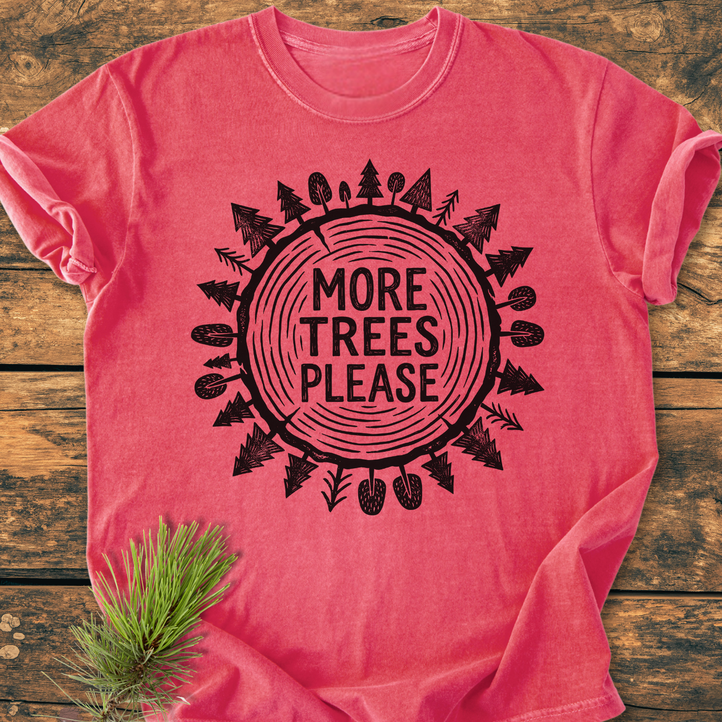 More Trees Please - Vintage Wash Tee features a circular tree-ring motif with surrounding tree illustrations and slogan, laid on wooden surface with a pine needle sprig, highlighting its woodland theme.