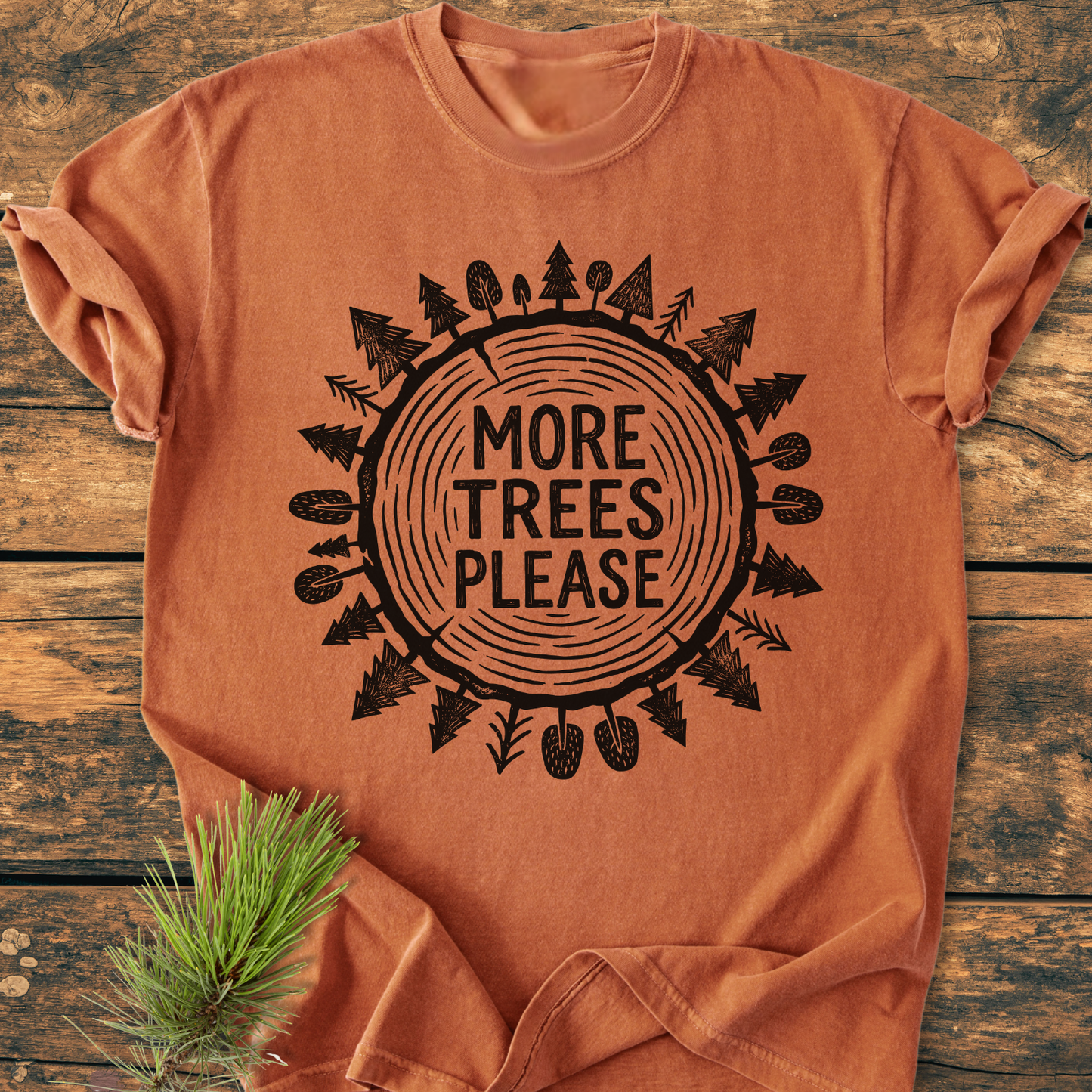 More Trees Please - Vintage Wash Tee features a tree trunk design with “MORE TREES PLEASE” text, laid flat on wood, with a pine sprig accenting its eco-friendly theme.