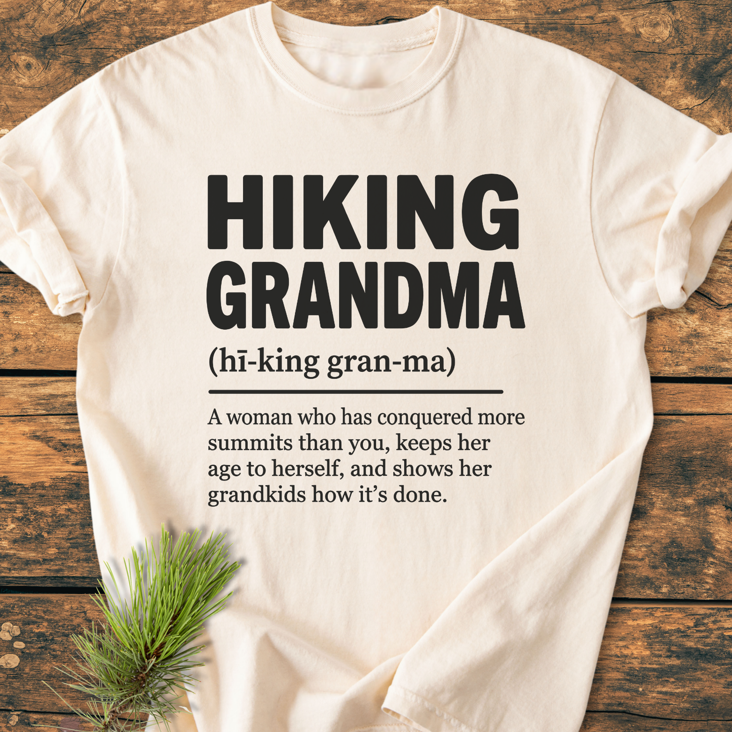 Cream-colored “Hiking Grandma Definition - Vintage Wash Tee” with bold black print, laid on wood. Features cheeky definition and pine sprig accent, highlighting outdoor theme. 100% ring-spun cotton, relaxed fit.
