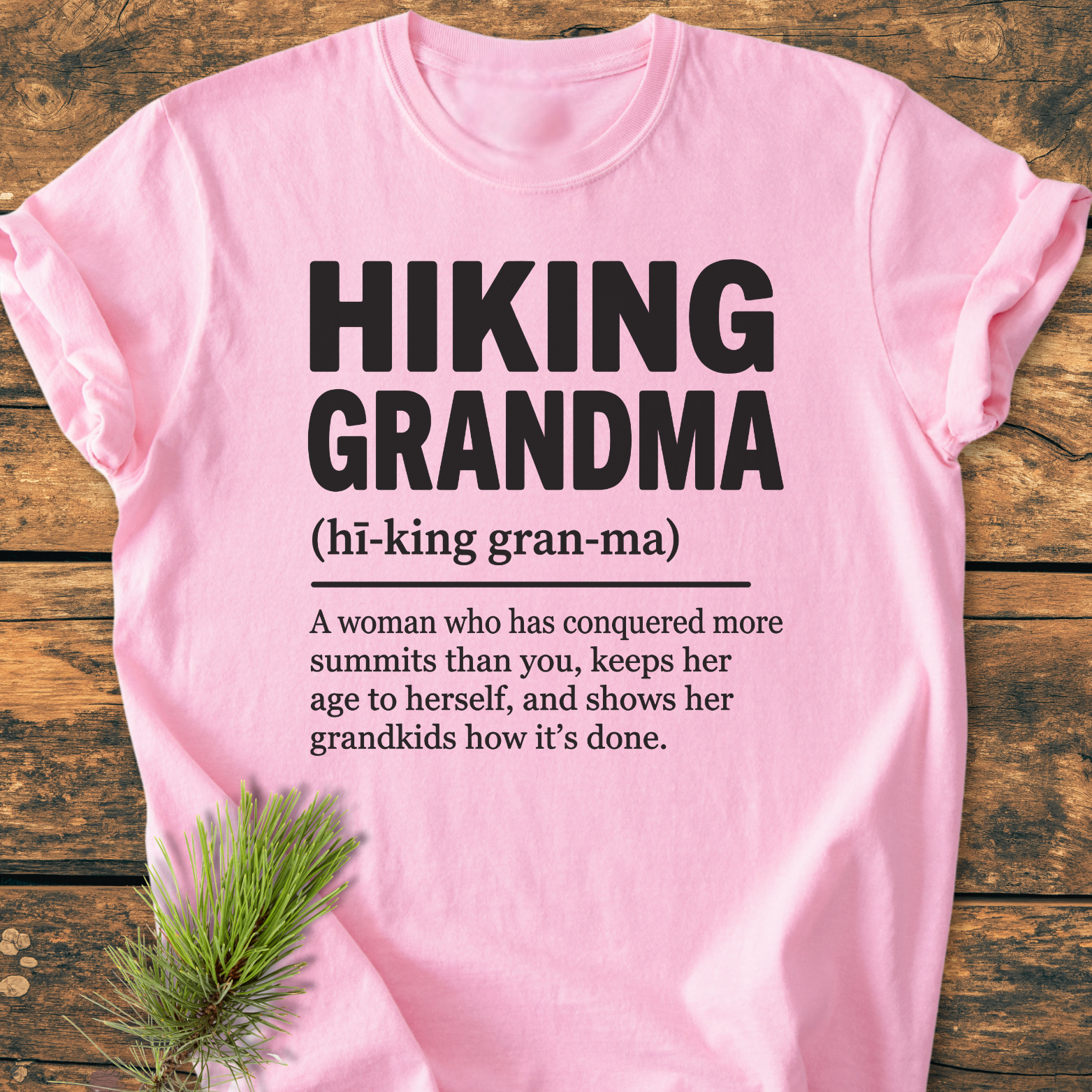 The Hiking Grandma Definition - Vintage Wash Tee features bold lettering and a playful definition, laid flat with pine needles on a wooden surface, showcasing its relaxed, durable design.