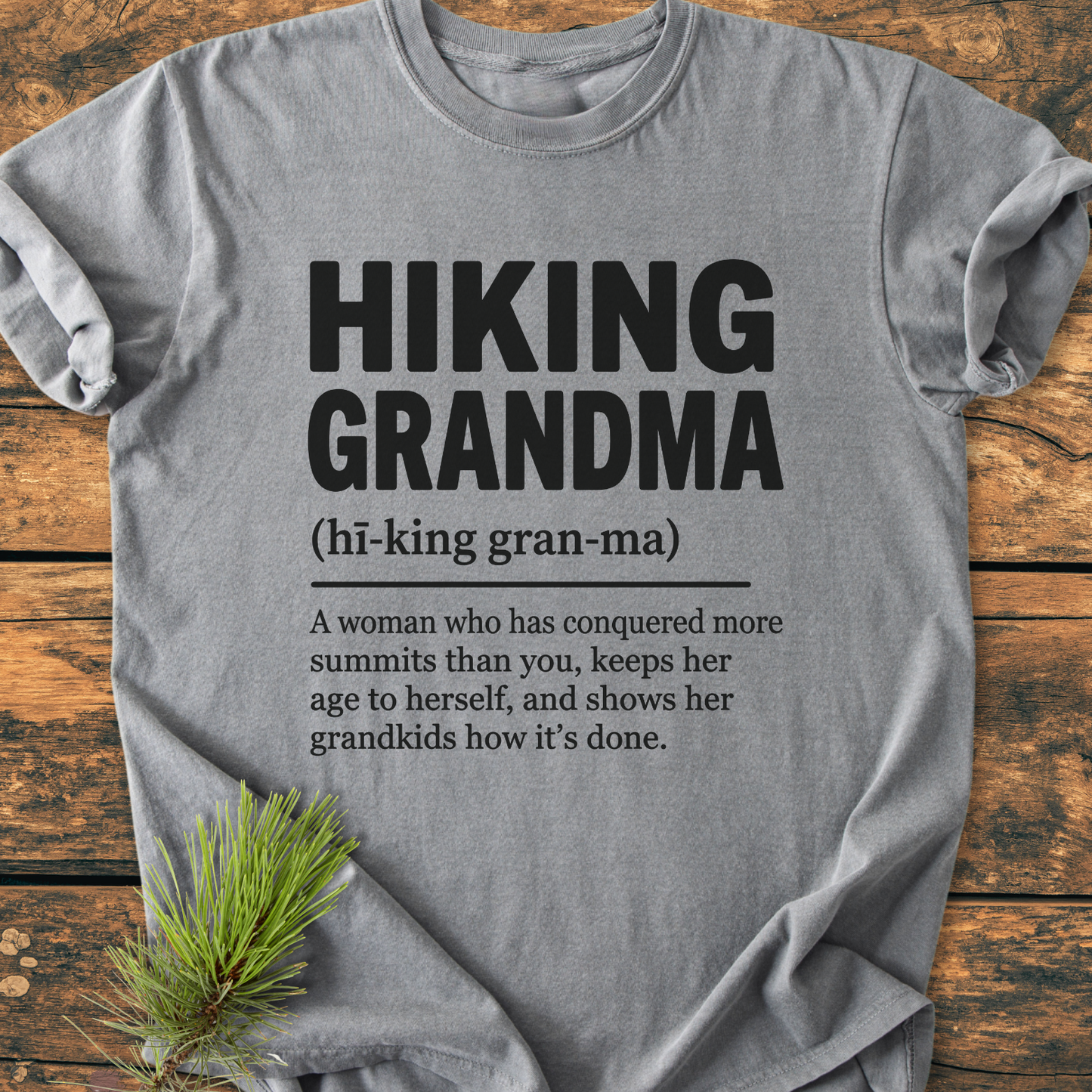Heather-gray Hiking Grandma Definition - Vintage Wash Tee with bold black text, phonetic spelling, humorous definition, on a wooden surface with a pine sprig prop for an outdoorsy touch.