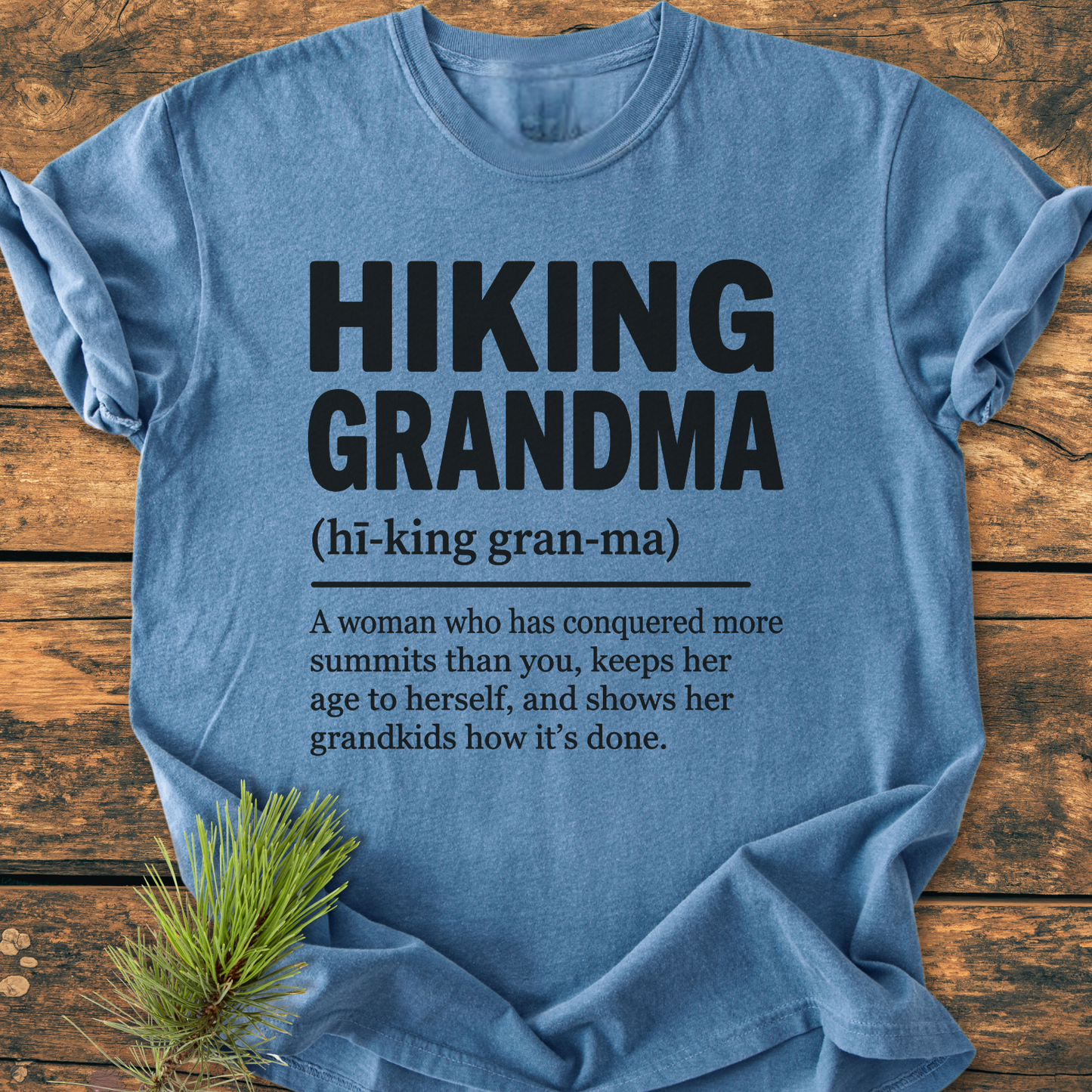 Heather-blue T-shirt featuring Hiking Grandma and dictionary-style definition, laid flat on wood with pine sprig. Soft, garment-dyed fabric, relaxed fit, highlighting vintage wash style.