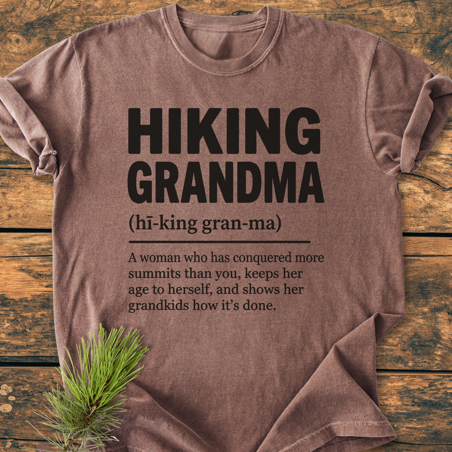Hiking Grandma Definition - Vintage Wash Tee: A brown, worn-look T-shirt with bold black text, laid flat on rustic wood, featuring a decorative pine sprig. Relaxed fit, pre-shrunk, 100% ring-spun cotton.