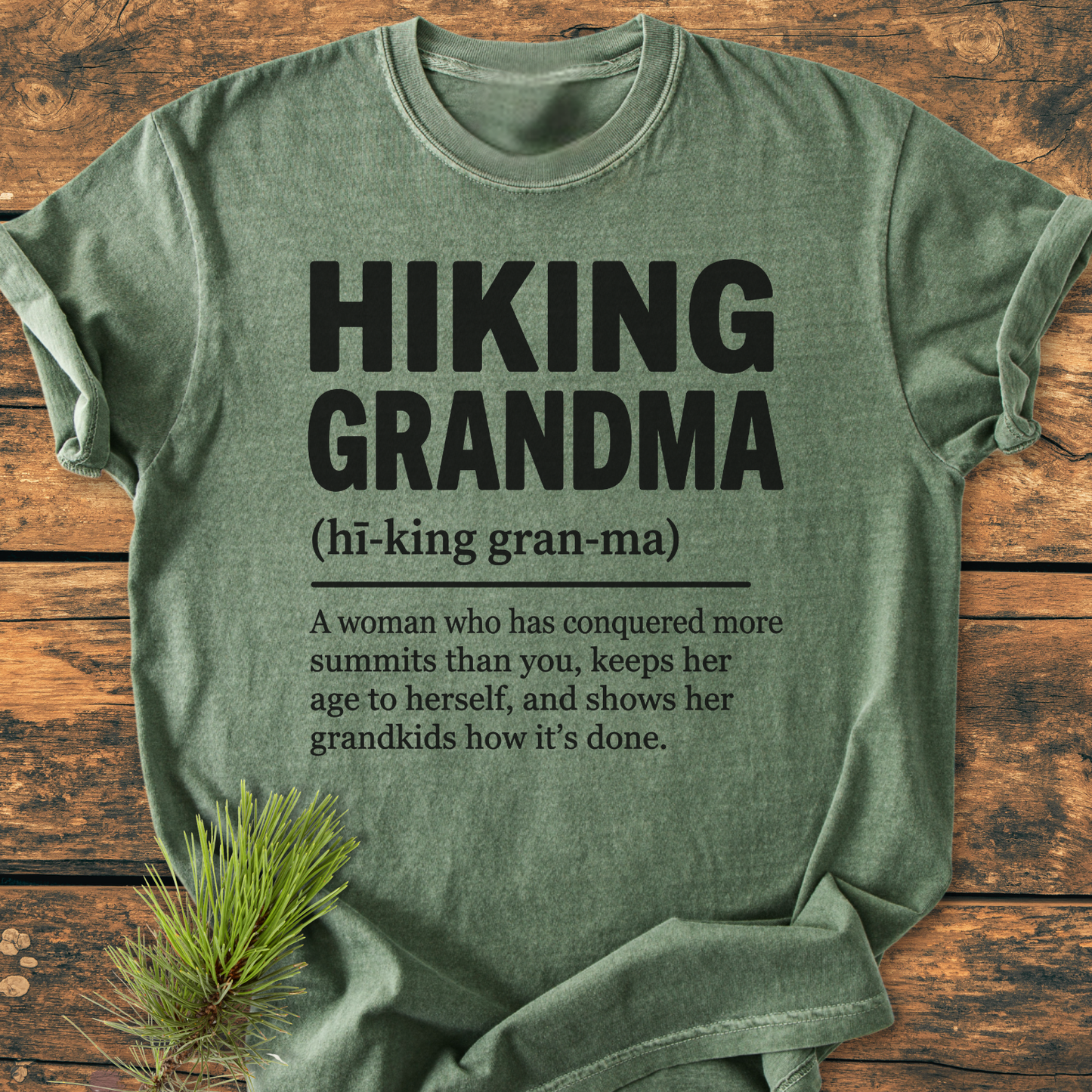 Hiking Grandma Definition - Vintage Wash Tee features bold black text on a green-heather T-shirt, laid out on a wooden surface, highlighting a playful definition celebrating adventurous grandmothers.