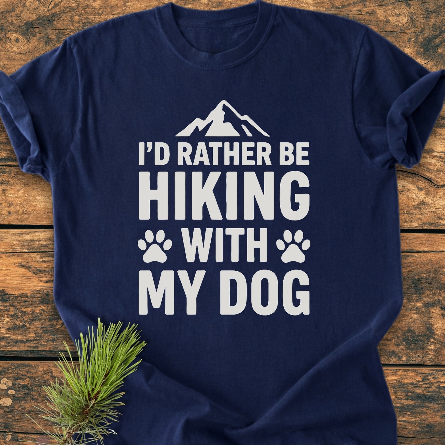 Navy-blue I'd Rather Be Hiking With My Dog - Vintage Wash Tee features bold white text, mountain silhouette, and paw prints. Laid flat with evergreen sprig, highlighting its relaxed fit and durable craftsmanship.