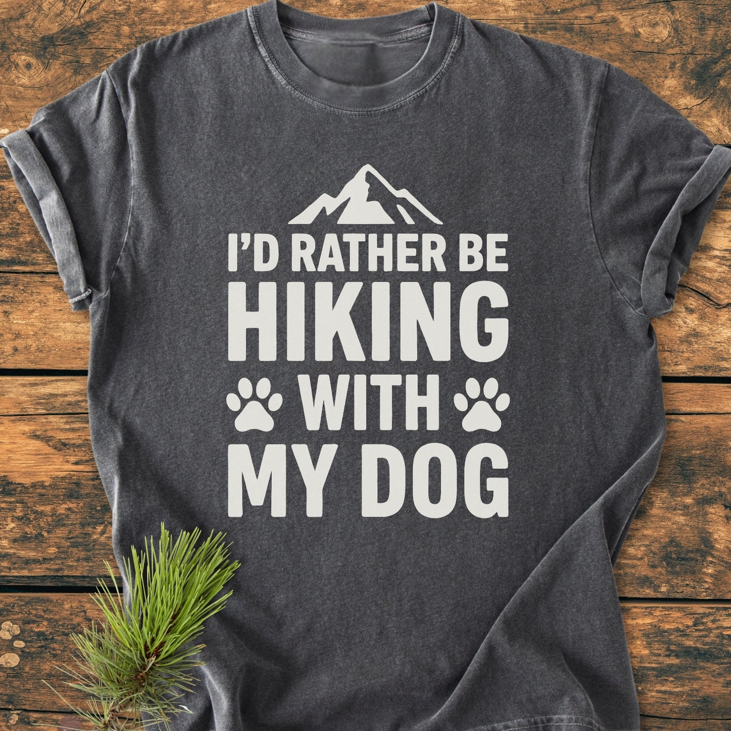Charcoal-gray I'd Rather Be Hiking With My Dog - Vintage Wash Tee displayed on wood with pine sprig, featuring white print, mountain silhouette, and paw prints, highlighting its cozy, relaxed fit and durable stitching.