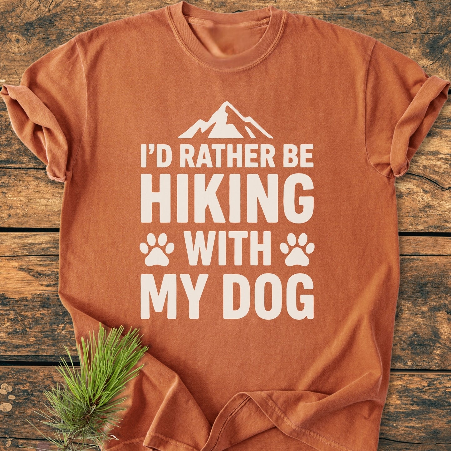 I'd Rather Be Hiking With My Dog - Vintage Wash Tee, featuring bold white text and mountain graphic on 100% ring-spun cotton, laid flat with rustic pine sprig accent on wooden background.
