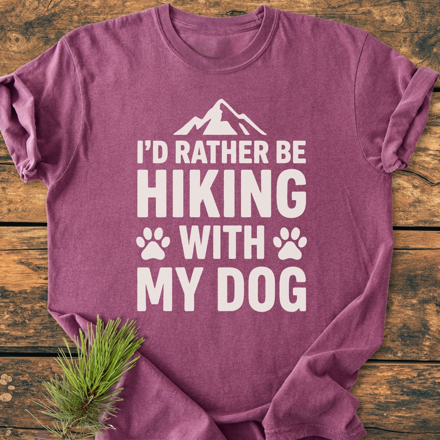 I'd Rather Be Hiking With My Dog - Vintage Wash Tee, plum-colored, folded on wood, features a mountain graphic, paw prints, and slogan in white. Pine sprig beside enhances outdoorsy theme.