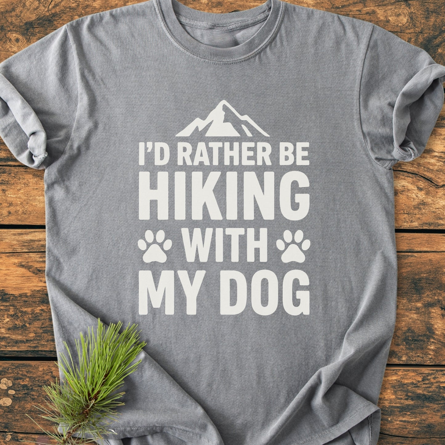 Heather-gray I'd Rather Be Hiking With My Dog - Vintage Wash Tee featuring a mountain icon and paw prints, laid flat on wood with a pine sprig beside it.