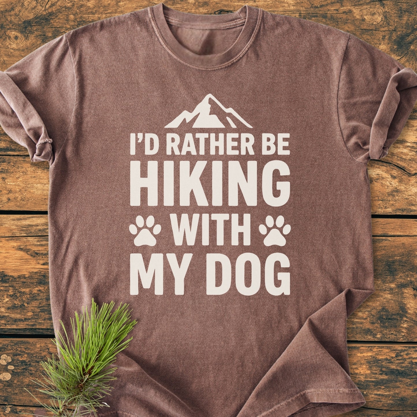 Brown I'd Rather Be Hiking With My Dog - Vintage Wash Tee displayed flat with white slogan and mountain icon, evoking an outdoorsy feel. Laid on wood, enhanced by an evergreen sprig.