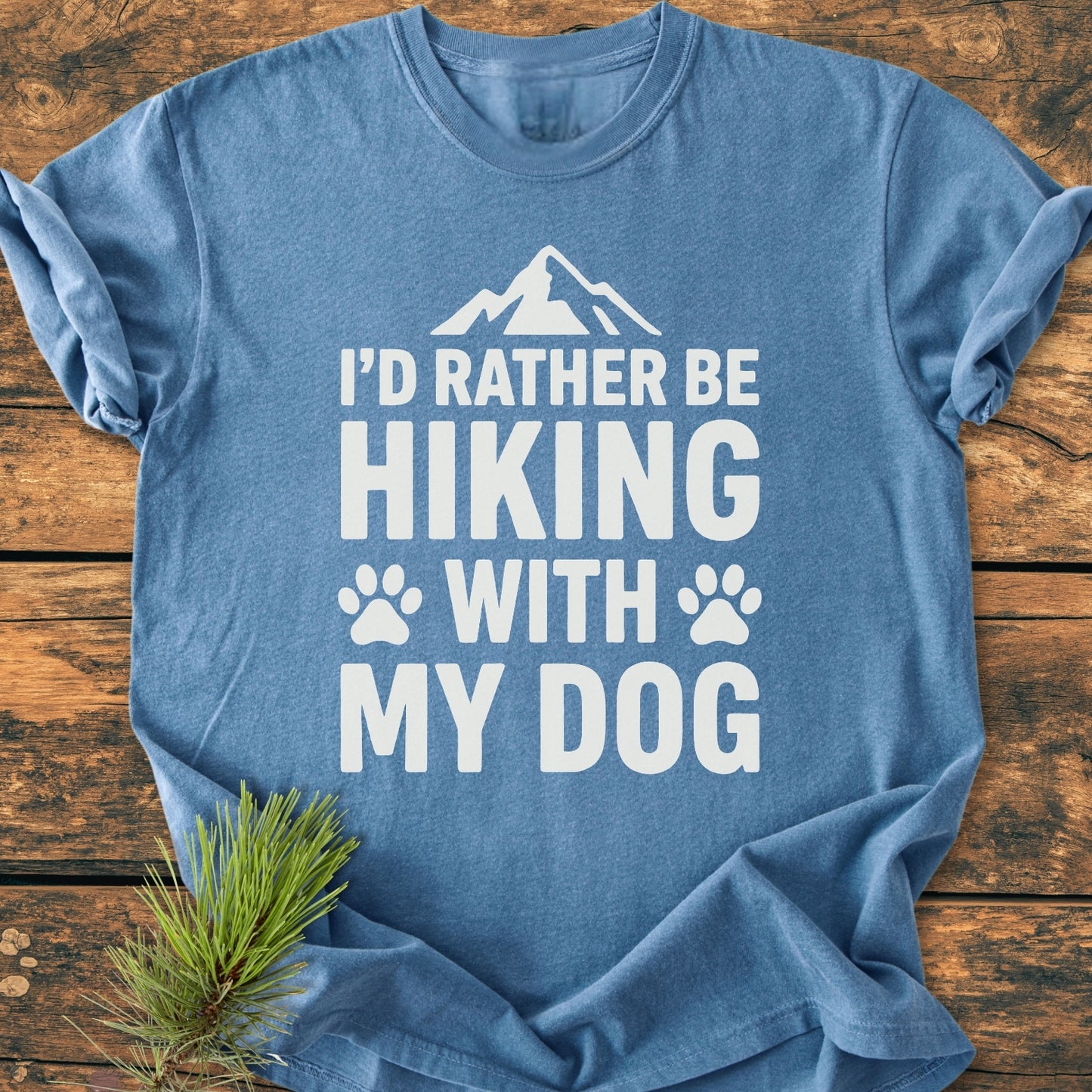 Heather-blue I'd Rather Be Hiking With My Dog - Vintage Wash Tee with white slogan and simple mountain graphic, laid flat with evergreen sprig beside it on wooden surface.