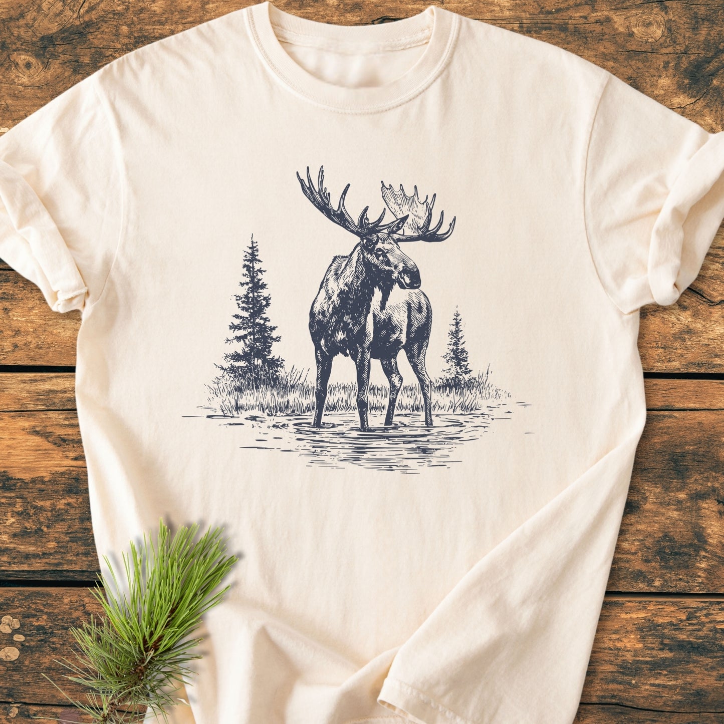 Moose - Vintage Wash Tee displayed flat on rustic wood, showcasing a detailed moose sketch with pine trees. Made from soft, pre-shrunk, garment-dyed cotton for comfort and durability.