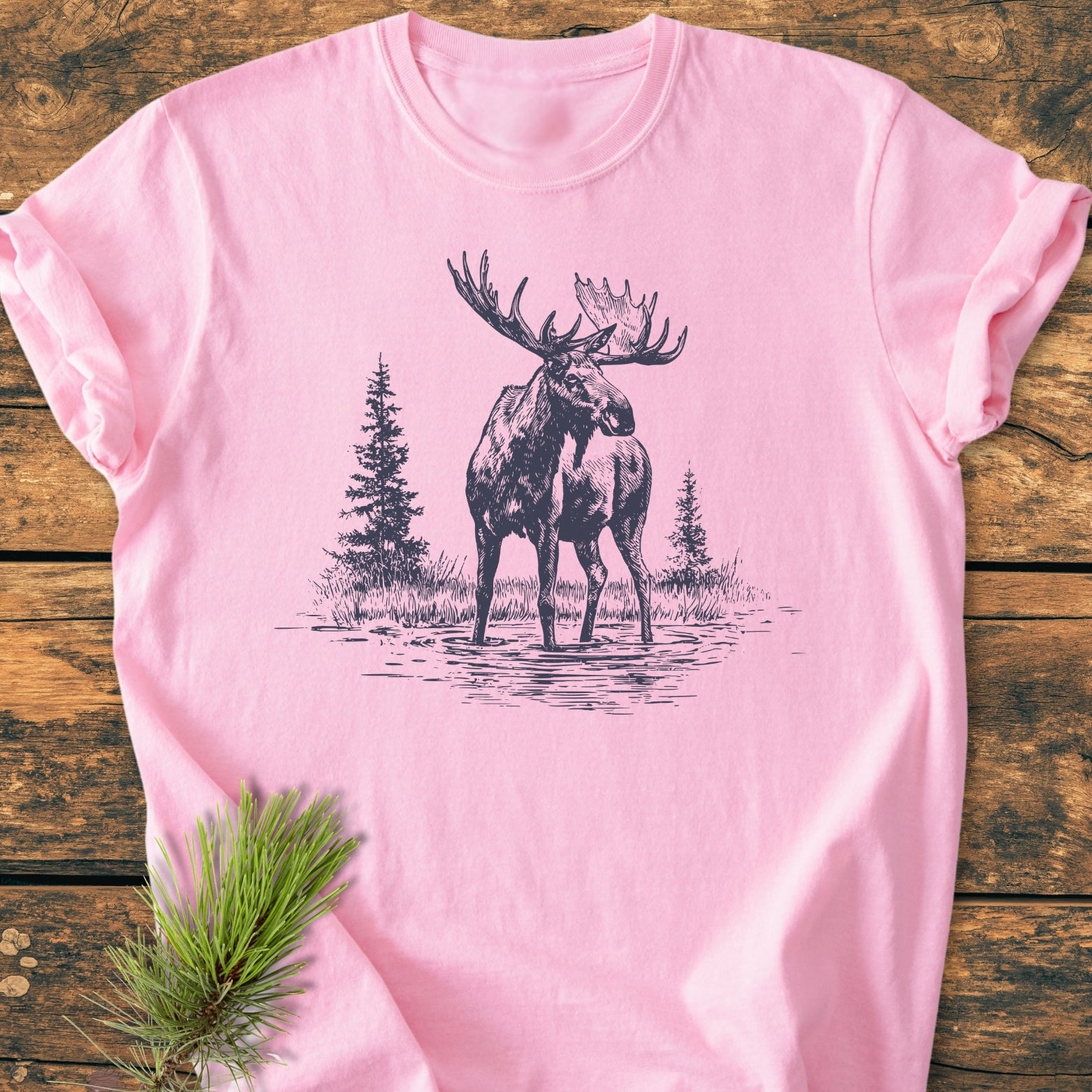 Moose - Vintage Wash Tee: A soft pink T-shirt featuring a sketch of a bull moose in water, with evergreen trees, laid on rustic wood. Includes a pine needle sprig accent.