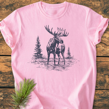 Moose - Vintage Wash Tee: A soft pink T-shirt featuring a sketch of a bull moose in water, with evergreen trees, laid on rustic wood. Includes a pine needle sprig accent.