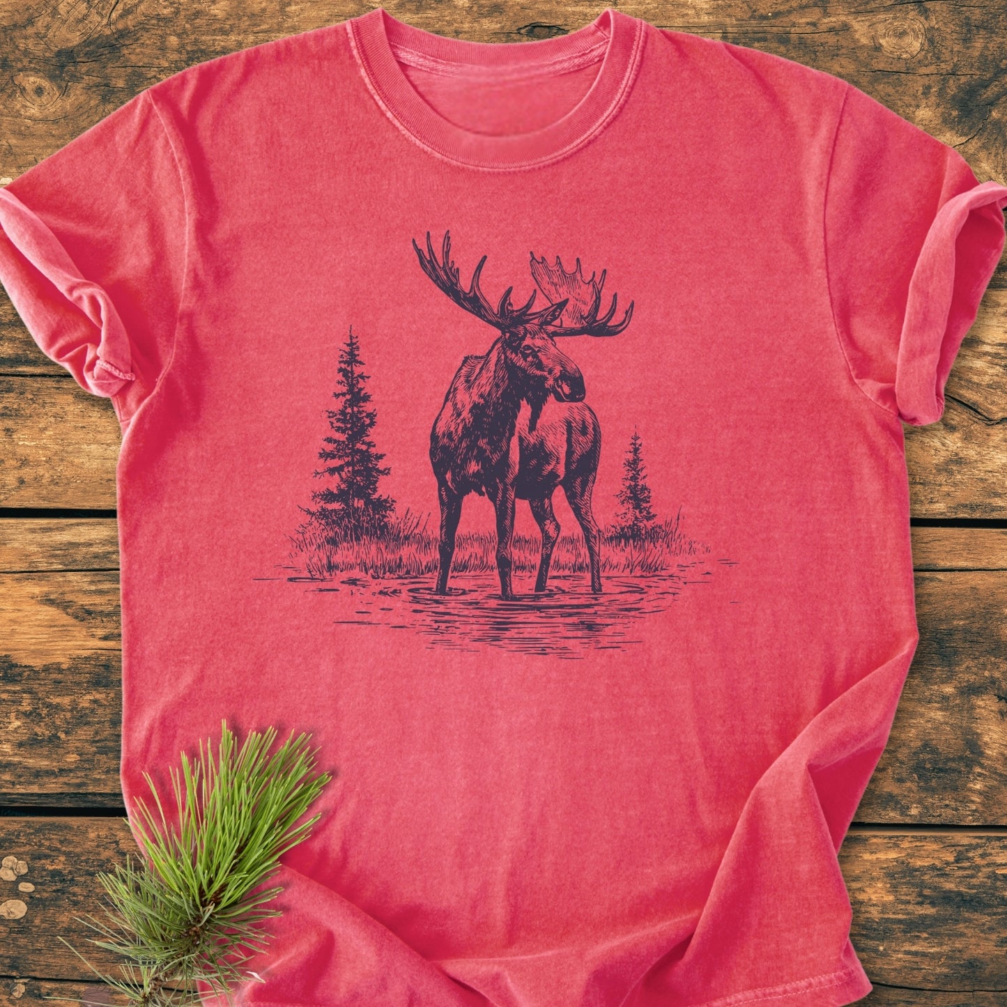 Moose - Vintage Wash Tee features a sketch-style moose print with pine trees on a faded red, 100% ring-spun cotton T-shirt, laid on wood, showcasing its relaxed fit and durable stitching.