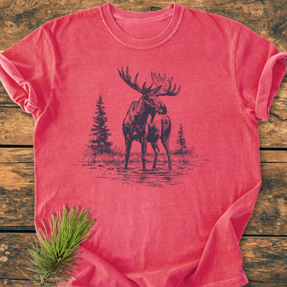 Moose - Vintage Wash Tee features a sketch-style moose print with pine trees on a faded red, 100% ring-spun cotton T-shirt, laid on wood, showcasing its relaxed fit and durable stitching.