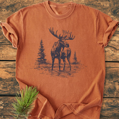 Moose - Vintage Wash Tee features a screen-printed bull moose design with evergreen trees on a rust-orange T-shirt, styled on a wooden backdrop with a pine sprig accent, offering a cozy, durable fit.