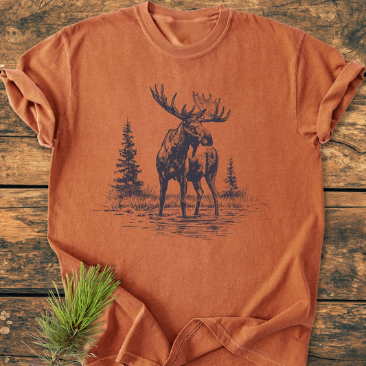 Moose - Vintage Wash Tee features a screen-printed bull moose design with evergreen trees on a rust-orange T-shirt, styled on a wooden backdrop with a pine sprig accent, offering a cozy, durable fit.