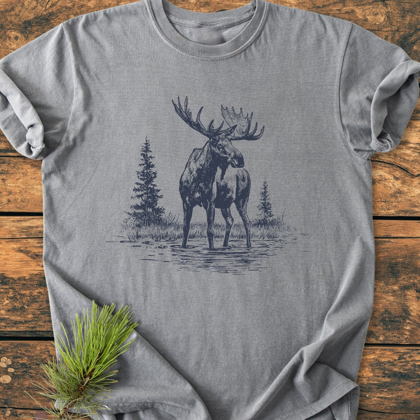 Moose - Vintage Wash Tee featuring a line-art bull moose design, laid on rustic wood. Soft-washed, 100% ring-spun cotton, relaxed fit, durable double-needle stitching, with a pine sprig accent.