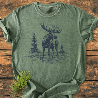 Moose - Vintage Wash Tee: A heather-green T-shirt featuring a printed wildlife scene of a bull moose in water with evergreen trees, laid flat on a wooden surface with a pine sprig nearby.