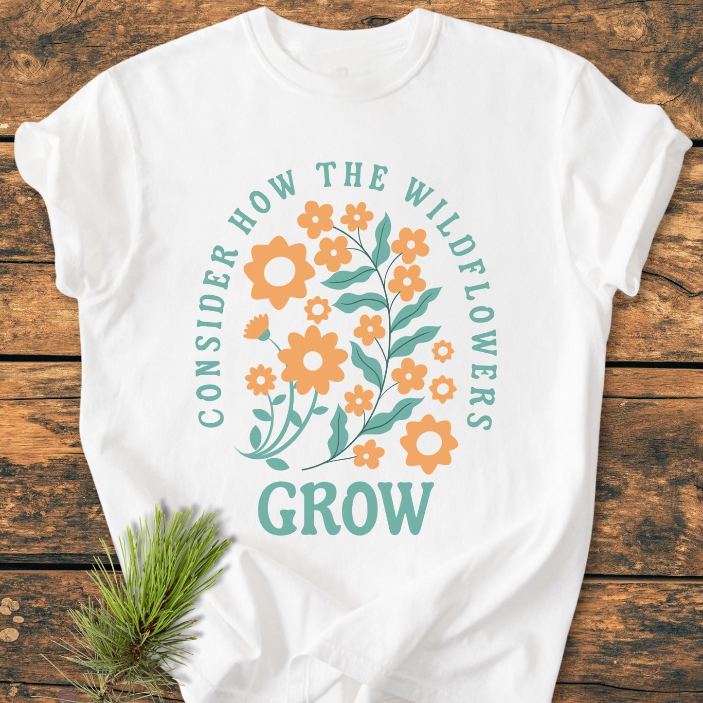 Wildflowers Luke 12,27 - Vintage Wash Tee featuring a floral graphic with CONSIDER HOW THE WILDFLOWERS GROW text on a white shirt, laid on a wooden surface with pine sprig.